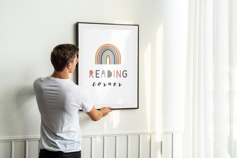 May include: A person hangs a framed print on a white wall. The print features a rainbow with the text "READING corner" in black lettering.