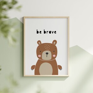 K&ouml;nnte beinhalten: Ein gerahmter Kunstdruck mit einer Cartoon-B&auml;ren-Illustration. Der B&auml;r ist braun mit einem beigen Bauch und rosa Wangen. Die Worte "be brave" sind in Schwarz &uuml;ber dem B&auml;ren gedruckt. Der Rahmen ist hellbraun.