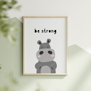 K&ouml;nnte beinhalten: Gerahmter Kunstdruck mit einer Cartoon-Nilpferd-Illustration in Graut&ouml;nen, mit rosa Akzenten und einem freundlichen Ausdruck. Die Worte "be strong" sind in Schwarz &uuml;ber dem Nilpferd gedruckt. Das Kunstwerk befindet sich vor einem wei&szlig;en Hintergrund.