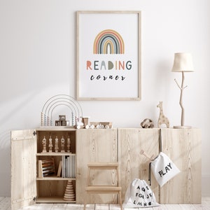 May include: A wooden cabinet with open doors, revealing shelves filled with toys and books. A rainbow-coloured arch with the text "READING corner" is displayed on the wall above the cabinet. There are two white canvas bags with the words "PLAY" and "ABC" on them.