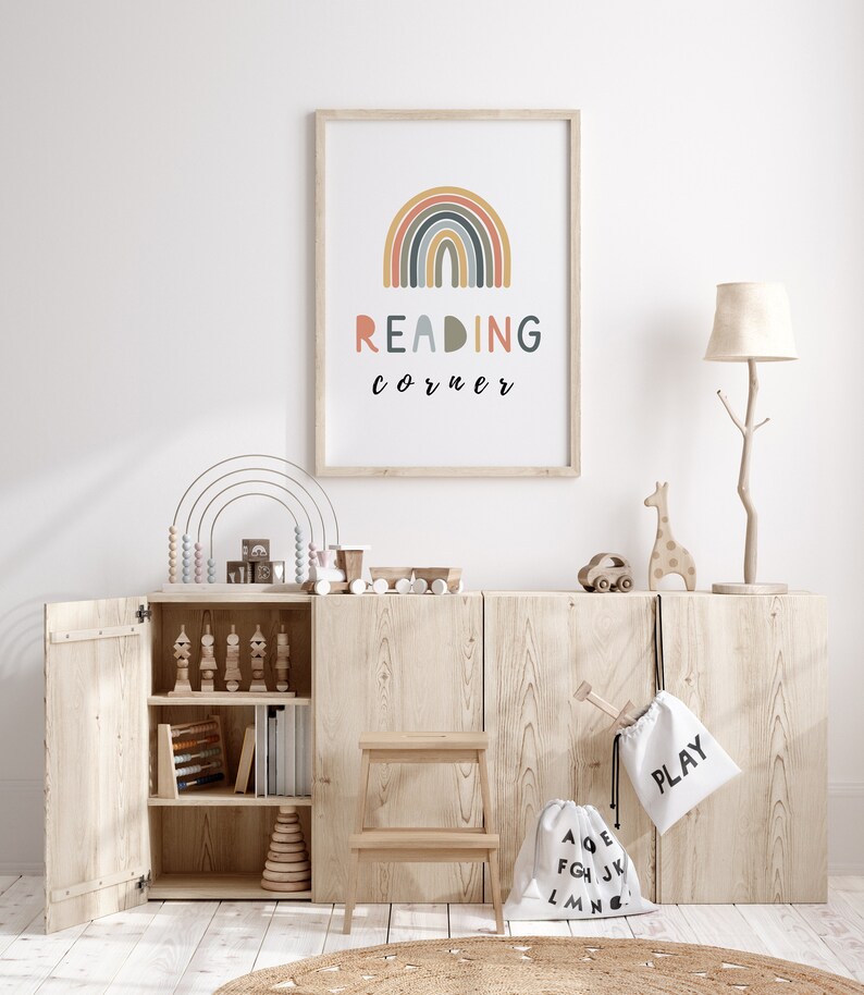 May include: A wooden cabinet with open doors, revealing shelves filled with toys and books. A rainbow-coloured arch with the text "READING corner" is displayed on the wall above the cabinet. There are two white canvas bags with the words "PLAY" and "ABC" on them.