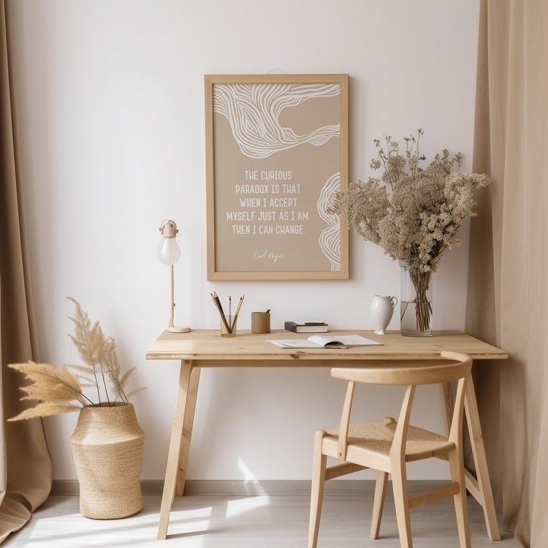 May include: A beige framed print with a white abstract design and the text "The curious paradox is that when I accept myself just as I am, then I can change." The print is hanging on a white wall above a wooden desk with a lamp, a vase of dried flowers, and a chair.