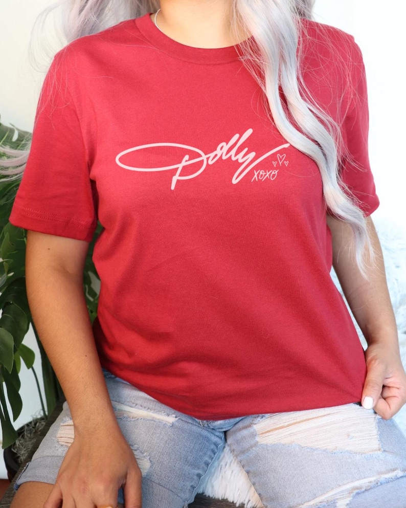 May include: A red short-sleeved t-shirt featuring the word Dolly in a white cursive script font across the chest. Below the name, the text xoxo is printed in a smaller white font, accompanied by three small white heart outlines.