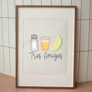 May include: A framed print with a white background and a simple illustration of a salt shaker, a shot glass with a yellow liquid, and a lime wedge. The text "Tres Amigos" is written below the illustration.