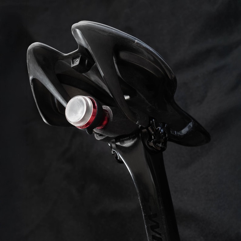 May include: A black bicycle seat with a small, white and red light attached to the back.