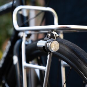 Peut inclure: Un porte-bagages de vélo en argent avec un feu de vélo noir fixé dessus. La lumière est allumée et éclaire la zone autour d'elle.