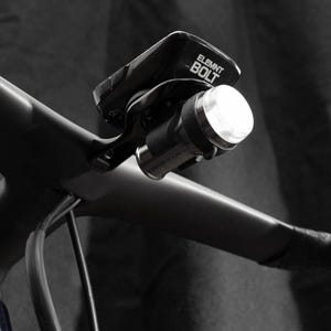 Puede incluir: Un ordenador de bicicleta negro con el texto "ELEMNT BOLT" montado en un manillar negro con una luz blanca adjunta.