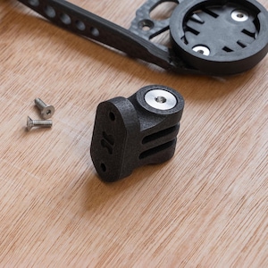 Peut inclure: Support de vélo en plastique noir avec trois fentes pour fixer des accessoires. Le support est fixé à une surface en bois avec deux vis argentées.