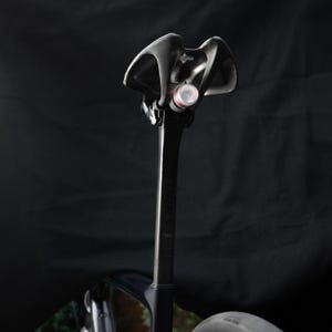 May include: Black bicycle seat post with a black and red bicycle light attached to the top.
