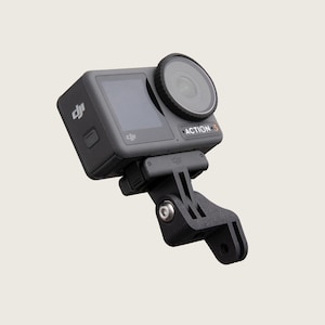 Puede incluir: Una cámara negra DJI Action 3 montada en un soporte negro estilo GoPro. La cámara tiene una pequeña pantalla en la parte posterior y una lente en la parte delantera. El soporte está inclinado hacia la derecha.