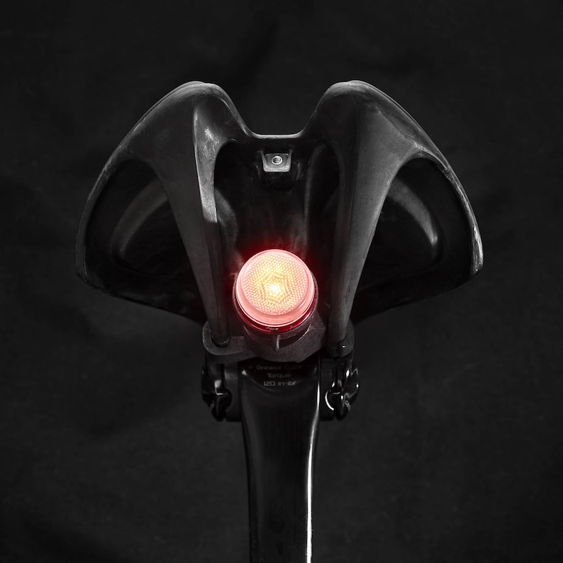 May include: A red bike tail light attached to a black bicycle seat post.