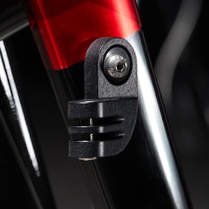 May include: Close-up of a black bicycle accessory attached to a red and black bike frame. The accessory has a bracket design with a screw and a mounting system. The bike frame is glossy and reflective.