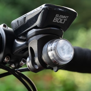 以下が含まれることがあります： 自転車のハンドルバーに取り付けられた黒いWahoo ELEMNT BOLTサイクルコンピューター。上部ケースには白いテキストロゴが目立ち、フロントマウントブラケットには半透明の白い円形のライトまたはアクセサリーが取り付けられています。