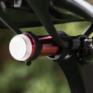 Puede incluir: Una luz de bicicleta roja con una lente blanca unida a un poste de asiento de bicicleta negro. La luz tiene el texto "EXPOSURE BOOSTR" impreso en ella.