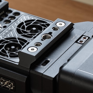 Peut inclure: Un appareil photo noir avec une plaque métallique sur le dessus. La plaque comporte quatre trous de vis et une découpe pour une vis. L'appareil photo est doté d'une batterie noire fixée au bas. Le texte "POWER TAP 3.0A MAX" est visible sur l'appareil photo.