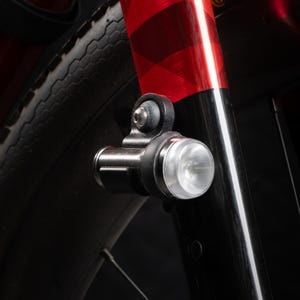 Peut inclure: Gros plan d'un phare de vélo noir monté sur la fourche avant. Le phare a une lentille claire et ronde et un corps noir. La fourche est noire et rouge avec un motif à carreaux. Une partie du pneu noir est visible.
