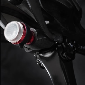 May include: A close-up of a black bicycle seat post with a red and white bike light attached. The light is mounted on the seat post with a black bracket. The light has a white lens and a red button.
