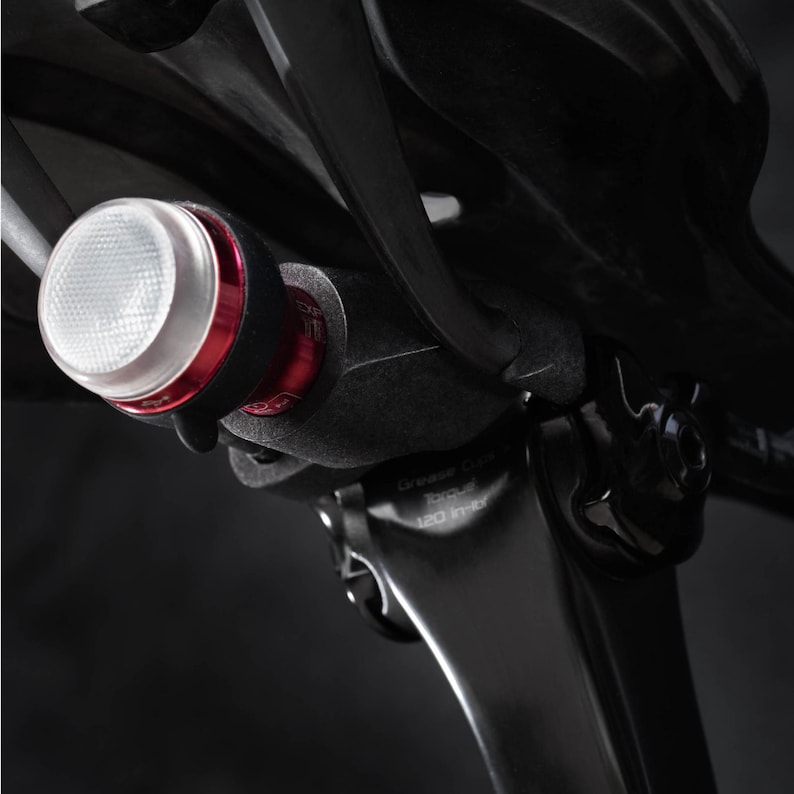 May include: A close-up of a black bicycle seat post with a red and white bike light attached. The light is mounted on the seat post with a black bracket. The light has a white lens and a red button.