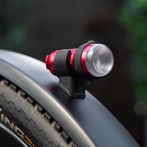 Peut inclure: Un feu de vélo rouge et noir est monté sur un garde-boue noir. La lampe a une lentille transparente et une sangle noire. Le pneu porte l'inscription "KING".