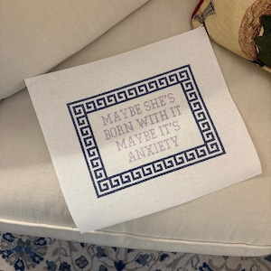 May include: A needlepoint canvas with the text "MAYBE SHE'S BORN WITH IT MAYBE IT'S ANXIETY" in lavender and navy blue. The text is framed by a navy blue Greek key border. The canvas is on a white surface.