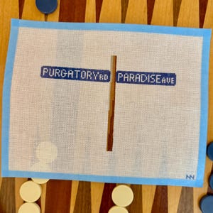 Könnte beinhalten: Nadelmalerei-Leinwand mit einem Straßenschild mit dem Text "Purgatory Rd" und "Paradise Ave" in blauer Schrift. Die Leinwand hat einen hellblauen Rand und ist von Backgammon-Steinen umgeben.