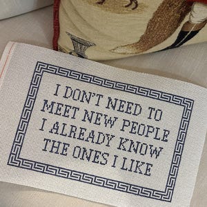Könnte beinhalten: Eine Gobelin-Leinwand mit dem Text "I DON'T NEED TO MEET NEW PEOPLE I ALREADY KNOW THE ONES I LIKE" in Dunkelblau. Der Text ist von einem dunkelblauen griechischen Schlüsselbordürenrahmen umgeben. Die Leinwand liegt auf einer hellbeigen Oberfläche.