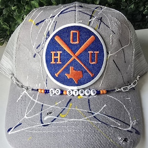 May include: Gray trucker hat with a patch featuring the letters "HOU" and crossed baseball bats, with a Texas outline. The hat has a beaded chain that spells "GO STROS" and is decorated with paint splatters.