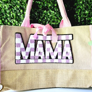 May include: A beige tote bag with pink trim and handles. The word "MAMA" is prominently displayed in a purple and white checkered pattern. A pink ribbon is tied at the top of the bag, adding a decorative touch.