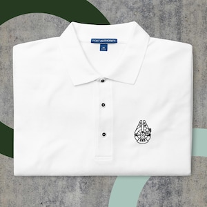 Può includere: Polo bianco con un'immagine ricamata nera del Millennium Falcon di Star Wars. La maglietta ha un'etichetta blu che dice "PORT AUTHORITY" e una taglia "M".