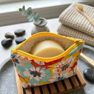 Peut inclure: Une petite pochette en tissu à fermeture éclair avec un motif floral vif composé de pétales rouges, jaunes et bleu clair. La pochette possède une fermeture éclair jaune vif et contient un pain de savon rond blanc cassé à l'intérieur.