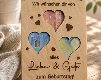 May include: A wooden birthday card with three heart-shaped cutouts revealing euro banknotes. The card has the German text "Wir wünschen dir von alles Liebe & Gute zum Geburtstag!" and small heart accents.