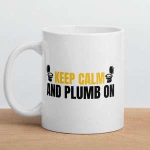 Puede incluir: Taza de cerámica blanca con un diseño en negro y amarillo. La taza presenta el texto "Keep Calm and Plumb On" con un icono de inodoro a cada lado del texto.
