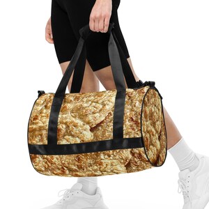 Pode incluir: Uma bolsa de viagem preta com um padrão marrom e dourado. A bolsa tem uma alça única e está sendo carregada por uma pessoa.