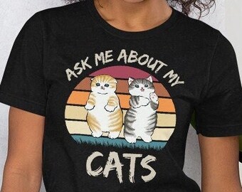 May include: A black t-shirt with a vintage-style graphic of two cats. The graphic features a sunset background with the text "Ask Me About My Cats".