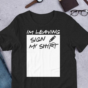 May include: Dark gray t-shirt with the text "IM LEAVING SIGN MY SHIRT" in black. The text is above a white rectangle with a pencil graphic. The shirt is displayed on a light gray surface with other accessories.