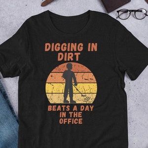 Könnte beinhalten: Schwarzes T-Shirt mit einer Grafik im Vintage-Stil einer Person, die einen Metalldetektor benutzt. Der Text lautet "Digging in Dirt Beats a Day in the Office."