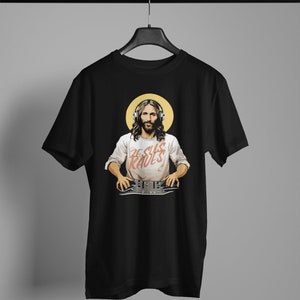 Peut inclure: T-shirt noir avec un motif de J&eacute;sus portant un casque et une chemise blanche avec le texte "Jesus Raves" imprim&eacute; dessus. Il tient une table de mixage DJ.