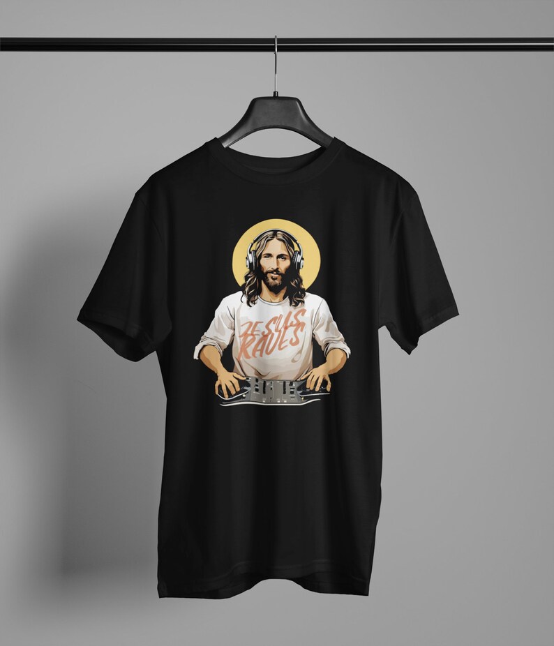 Peut inclure: T-shirt noir avec un motif de J&eacute;sus portant un casque et une chemise blanche avec le texte "Jesus Raves" imprim&eacute; dessus. Il tient une table de mixage DJ.