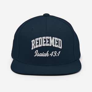 Puede incluir: Gorra de béisbol azul marino con visera plana. La palabra "REDEEMED" está bordada en blanco en la parte delantera, en forma de arco. Debajo, el texto "Isaiah 43:1" también está bordado en blanco. La gorra tiene una corona estructurada.