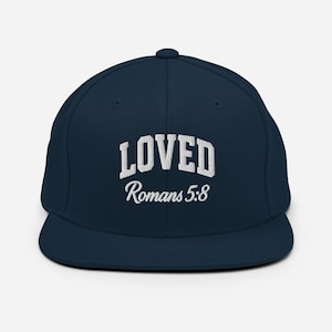 Puede incluir: Gorra de béisbol azul marino con la palabra "LOVED" bordada en blanco y "Romanos 5:8" debajo. La gorra tiene una visera plana y un diseño estructurado, ideal para un look casual.