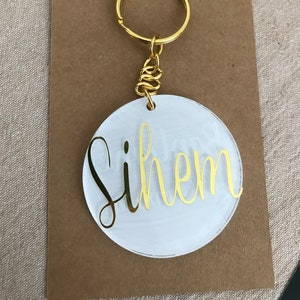 May include: A circular white acrylic keychain featuring the name Sihem in gold script lettering. The keychain is attached to a gold metal split ring by a short gold chain, presented on a rectangular brown cardboard display card with a top cutout.