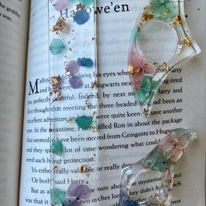 Peut inclure: Marque-page et bague en résine transparente ornés de fleurs séchées bleues, violettes et vertes, avec des accents de feuilles d'or. Le marque-page porte l'inscription "Chapter Ten Hallowe'en". La bague est conçue pour être portée au doigt.