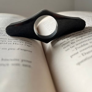 Peut inclure: Un marque-page noir en forme de boucle avec une ouverture circulaire au centre. Le marque-page est posé sur un livre ouvert.