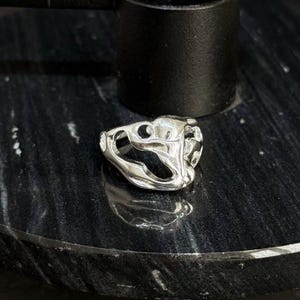 Könnte beinhalten: Ein silberfarbener Ring mit abstraktem Design, der offene Räume und geschwungene Linien aufweist. Der Ring wird auf einer schwarzen Marmoroberfläche neben einem schwarzen zylindrischen Objekt präsentiert.