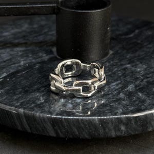 Könnte beinhalten: Ein silberner Ring mit Kettengliedern. Der Ring besteht aus ineinandergreifenden rechteckigen Gliedern. Er liegt auf einer dunkelgrauen und schwarzen Marmoroberfläche. Im Hintergrund befindet sich ein schwarzes Metallobjekt.