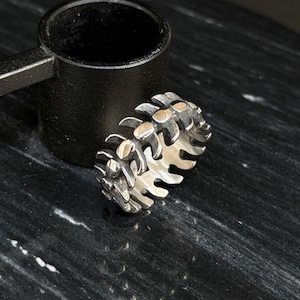 Könnte beinhalten: Ein silberfarbener Ring mit einem einzigartigen, strukturierten Design. Der Ring weist eine Reihe von miteinander verbundenen, stilisierten knochenartigen Formen auf. Der Ring liegt auf einer dunklen, marmorierten Oberfläche neben einem schwarzen Objekt.
