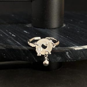 Könnte beinhalten: Ein verstellbarer Ring in Silberoptik mit einem kreisförmigen, cremefarbenen Mittelstück. Der Ring hat ein detailliertes Design auf dem Mittelstück und eine kleine, hängende Kugel. Der Ring wird auf einer dunklen, strukturierten Oberfläche präsentiert.