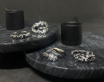 May include: Four silver rings with black stones on a black marble surface. The rings have a textured, organic design.