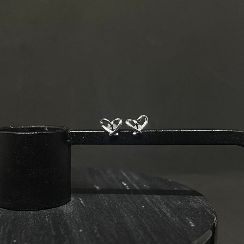 May include: A pair of silver heart-shaped stud earrings. The earrings are made of a single piece of wire that is twisted into a heart shape.