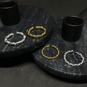 Könnte beinhalten: Drei gedrehte Metallringe in Silber, Gold und dunklerem Silber. Die Ringe werden auf einer schwarzen Marmorfläche präsentiert.